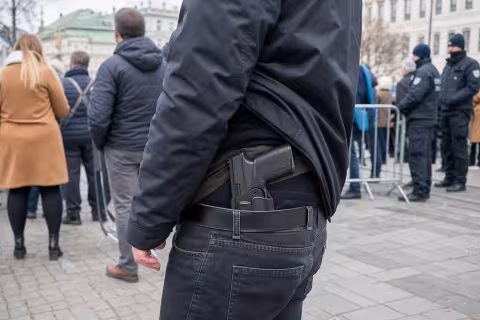 Skryto nosená pištoľ pod bundou na okraji verejného zhromaždenia s políciou v pozadí.